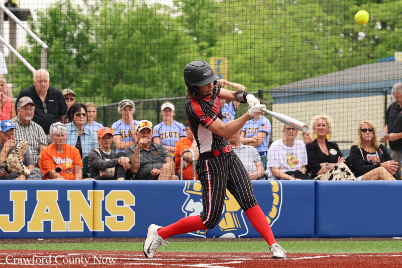 Female softball player in a black-and-white striped uniform swings at a pitch, wearing a helmet, with fans watching from the stands.