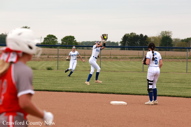 Softball game in progress: a fielder leaps to catch a high ball near second base while a teammate watches and a catcher in red is blurred in the foreground.