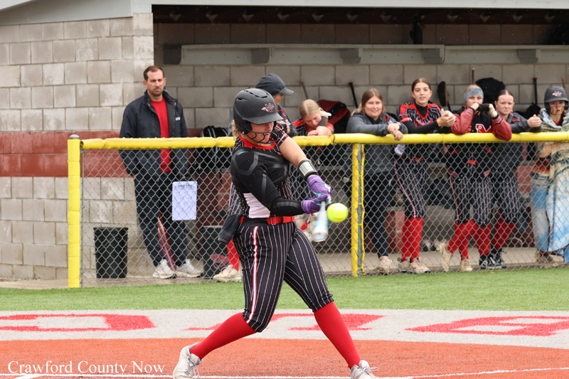 Softball batter swinging at a pitch, wearing a black-and-white uniform with red socks, as spectators watch from behind a chain-link fence behind home plate.