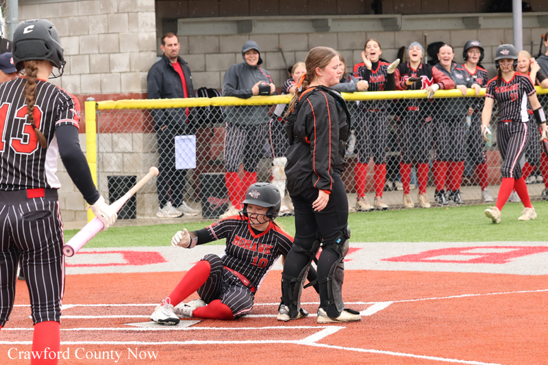 Softball catcher in black and red sits on the ground at home plate as a teammate in protective gear stands nearby, with fans cheering behind a chain-link fence.