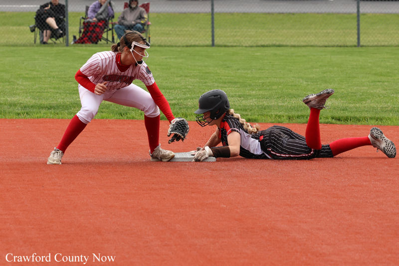 Softball baseman in white guards the bag as a runner in black slides headfirst into a base on an orange dirt infield, with spectators in the background.