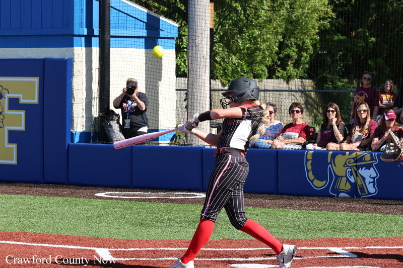 Female softball batter in black pinstripe uniform swings at a pitch, red socks, helmet on, on a turf field with fans watching behind the fence.