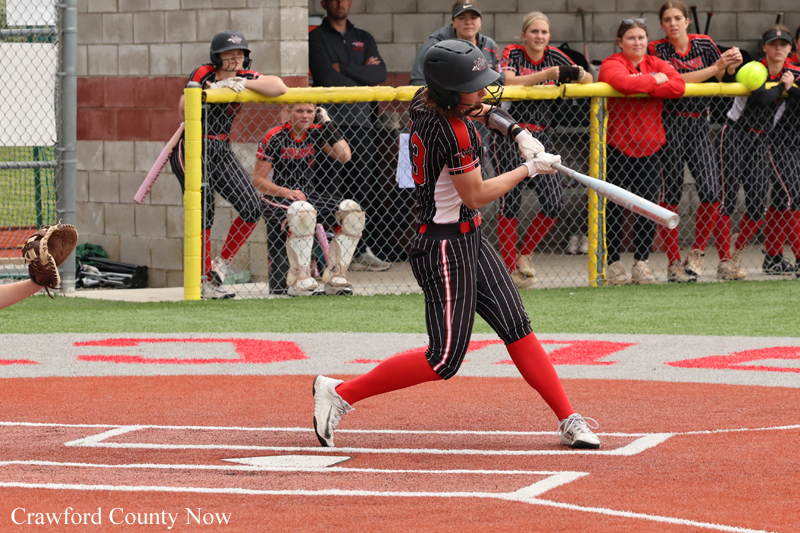 Female softball player in black-and-red uniform swings a bat at a pitch at home plate, crowd watching behind a chain-link fence.