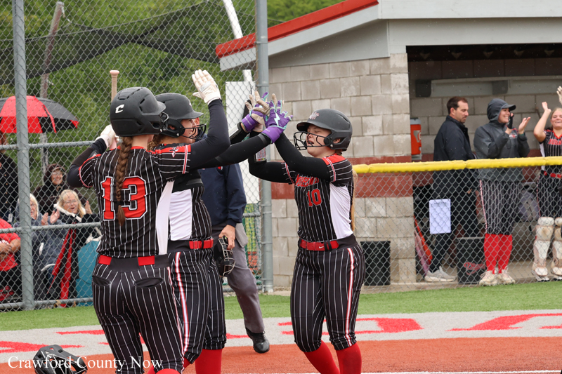 Softball players in black pinstriped uniforms high-five at home plate after a run as spectators watch from behind the fence.