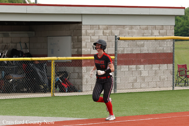 Female softball player in black and red uniform runs along the baseline wearing a helmet and batting gloves near a dugout fence. (Crawford County Now)
