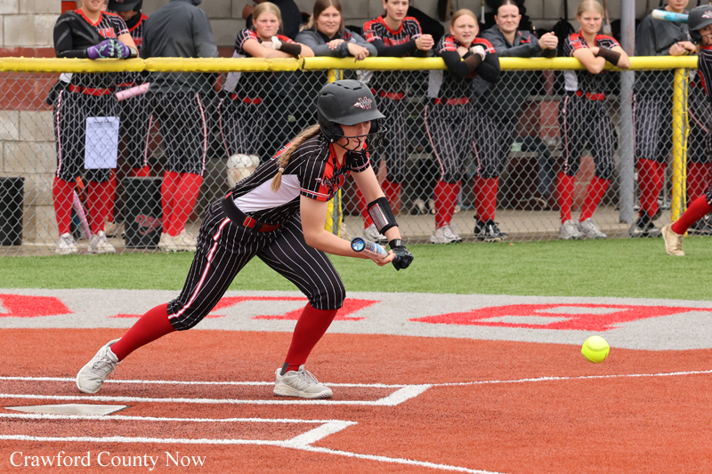 Female softball player in a black-and-red uniform sprints toward home plate as a ball approaches, with spectators watching behind a chain-link fence.