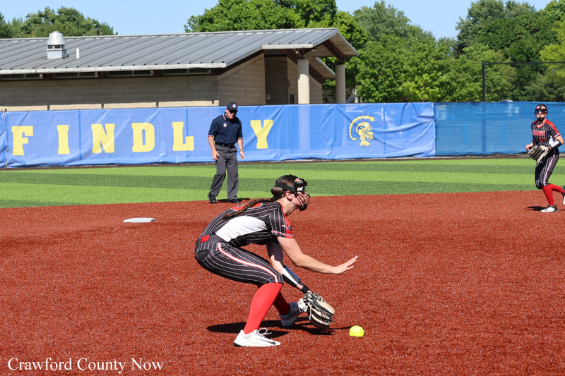 Softball infielder in a black-red uniform crouches to field a ground ball on a red dirt infield, with a yellow ball near her glove.