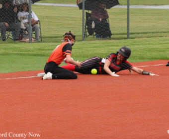 Softball game moment: a baserunner in a black and red uniform slides into a base as a teammate in an orange jersey kneels nearby with the ball on the dirt.