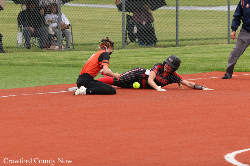 Softball game moment: a baserunner in a black and red uniform slides into a base as a teammate in an orange jersey kneels nearby with the ball on the dirt.