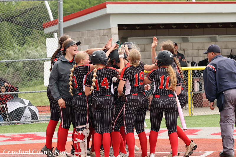 Youth girls' softball team huddling in the dugout, raising hands in celebration during a game.