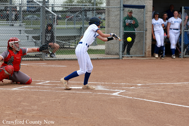 Female softball batter in a white uniform swings at a pitch as the ball approaches; catcher in red gear crouches behind home plate on a dirt field.