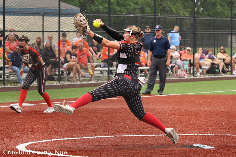 Softball pitcher in a black pinstriped uniform delivering a pitch on a red infield, with spectators behind a chain-link fence.