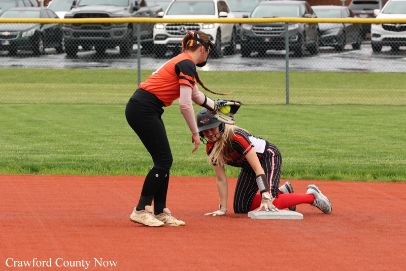 Coach guiding a young baserunner kneeling at first base during a softball game on a red infield.