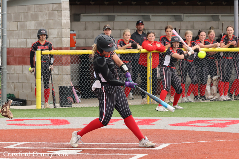 Female softball batter in black protective gear swings at a pitched ball near home plate, with teammates watching behind the chain-link fence.