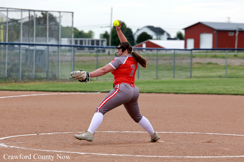 Right-handed softball pitcher in red and gray uniform mid-throw on a dirt mound at a field touchline close by a fence.