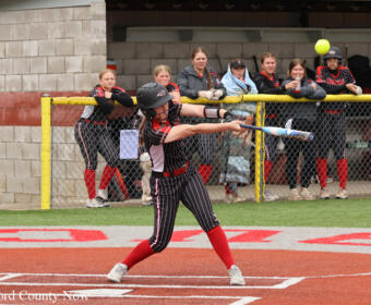 Female softball batter in a black pinstripe uniform with red socks swings at a pitch, as teammates and spectators watch from behind the fence in the dugout area.