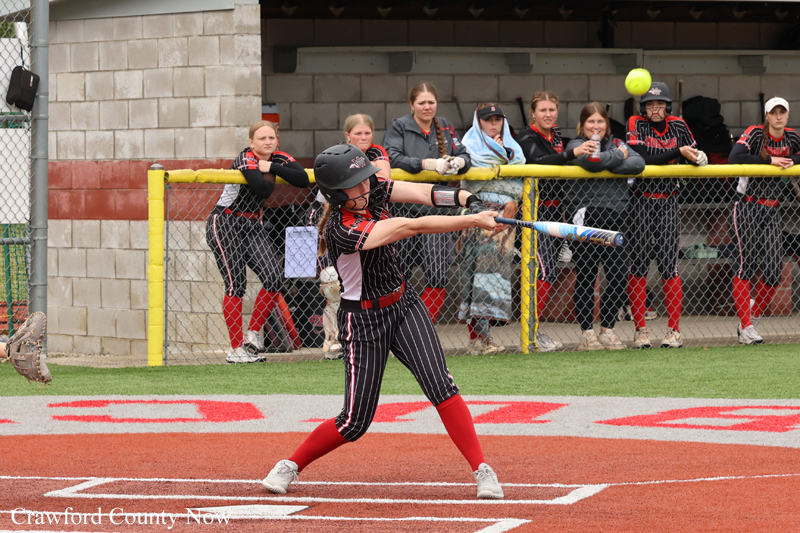Female softball batter in a black pinstripe uniform with red socks swings at a pitch, as teammates and spectators watch from behind the fence in the dugout area.