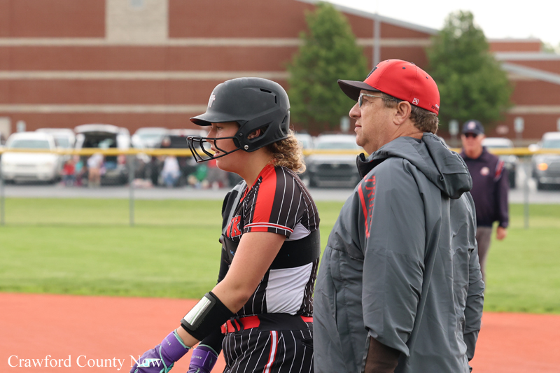 Softball player in a black-red uniform wearing a helmet and wrist guards stands with a coach on a dirt field with a school building in the background.