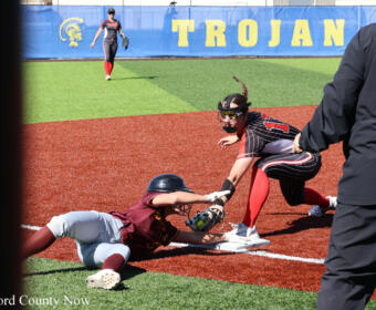Baserunner slides into a base as the catcher reaches to tag, while a third player watches; a blue wall with 'TROJAN' lettering is in the background.