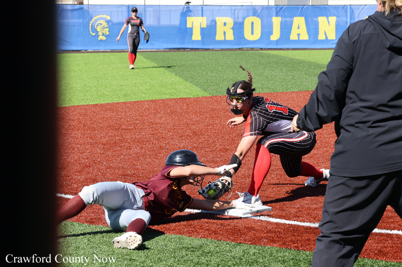 Baserunner slides into a base as the catcher reaches to tag, while a third player watches; a blue wall with 'TROJAN' lettering is in the background.