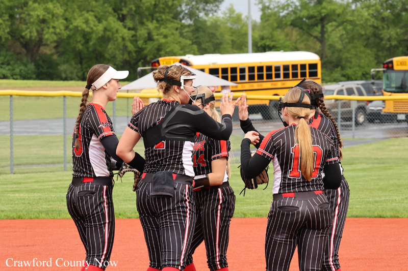 Softball players in black pinstripe uniforms high-five each other on the field, with two yellow school buses and a chain-link fence in the background.