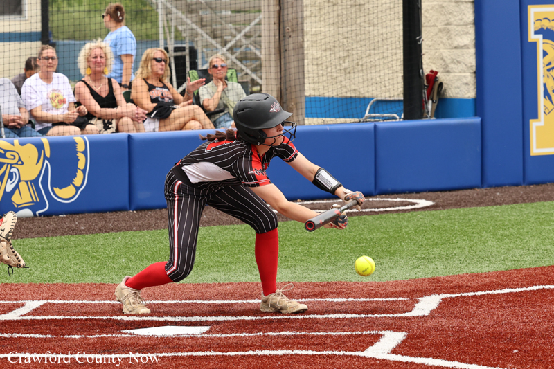 Female softball batter in pinstripe uniform and helmet reaches to swing at a yellow ball on a red dirt field as spectators watch from the blue outfield wall behind her.