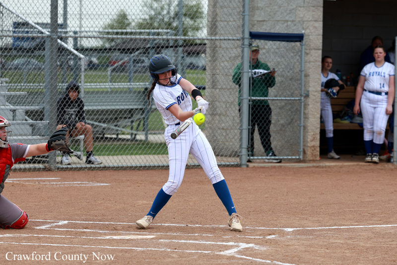 Young female softball batter swings at a pitch at home plate with catcher ready behind her and dugout in the background.