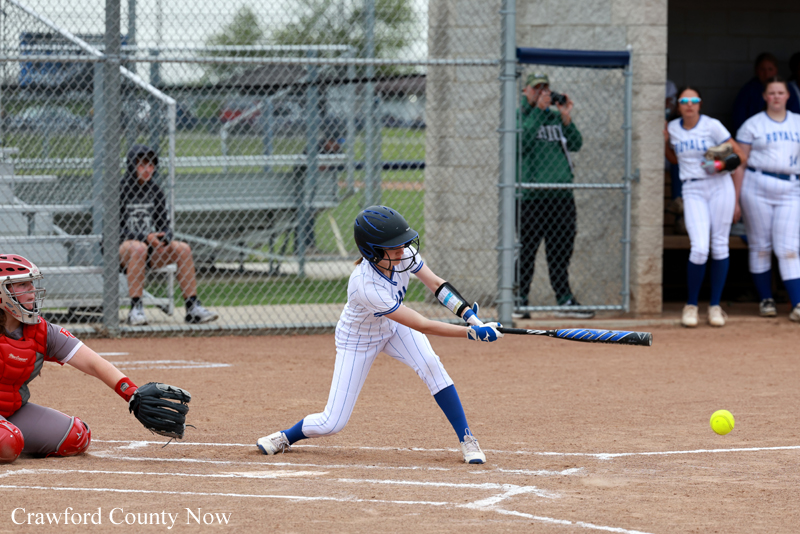 Young girl in a white uniform and blue helmet swings at a pitched ball; catcher in red crouches behind home plate with teammates watching from the dugout.