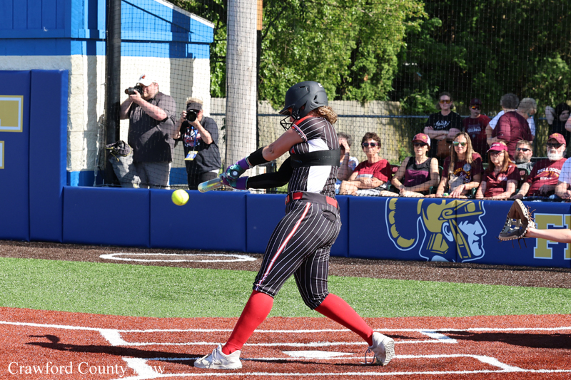 Female softball batter in striped uniform swings at a ball near home plate as photographers and spectators watch from the stands.