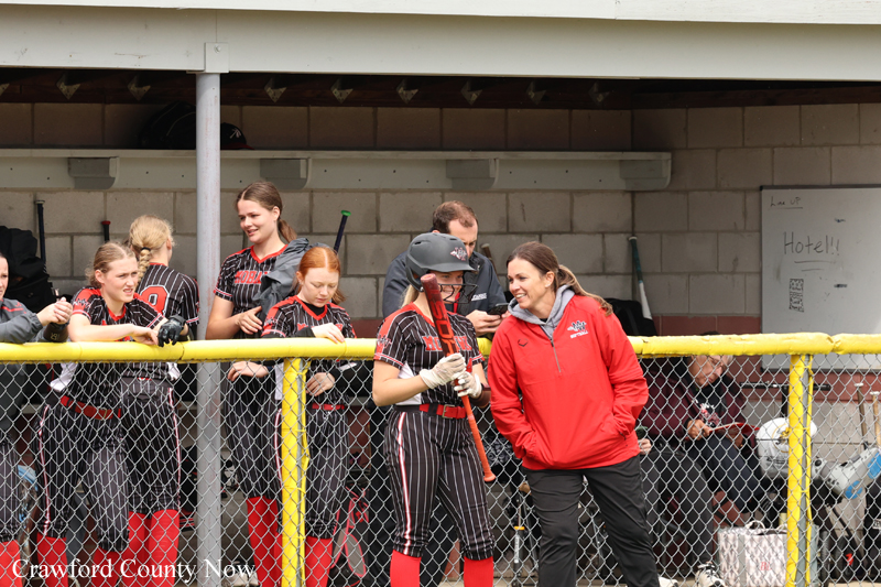 Girls' softball players in black-and-red uniforms lean on a yellow fence in the dugout, chatting with a coach in a red jacket.