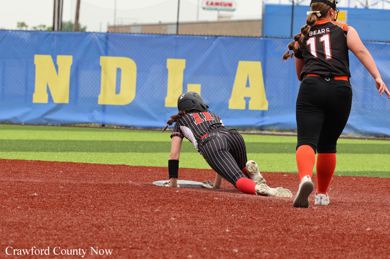 Softball baserunner slides headfirst toward a base as a teammate stands nearby on a red dirt infield with a blue outfield wall behind him or her.