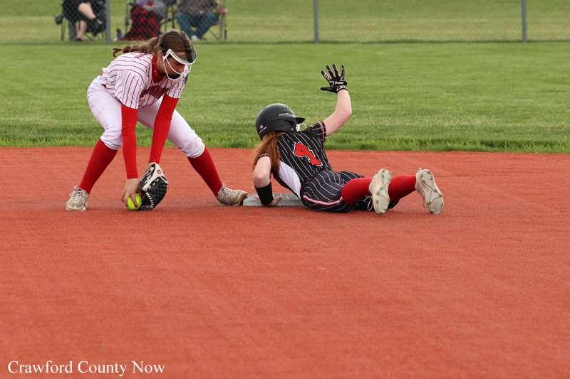 Two girls playing softball race for a ground ball on the red infield; one in white and red reaches with a glove, the other in a black pinstripe uniform slides into the base.