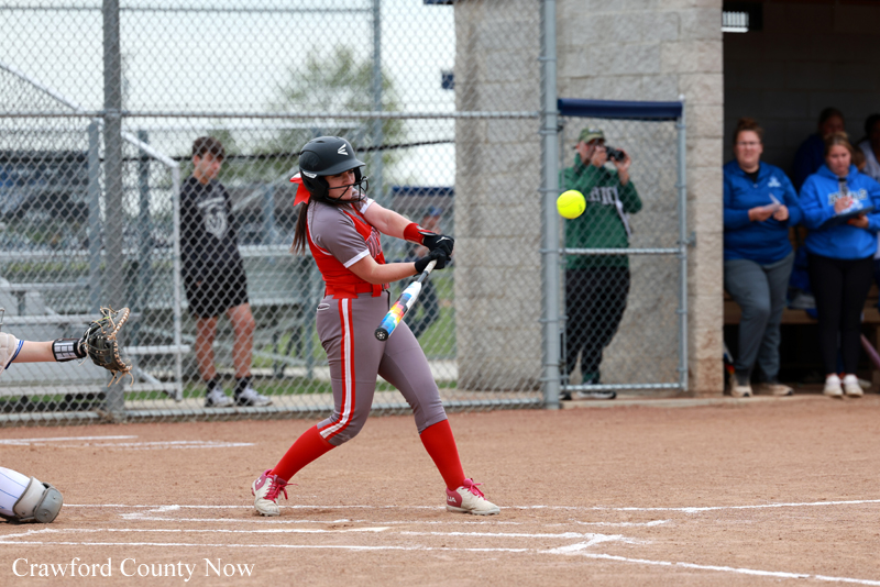 Female softball player in red hits the ball during a game; catcher crouches behind her and spectators watch from the fence.