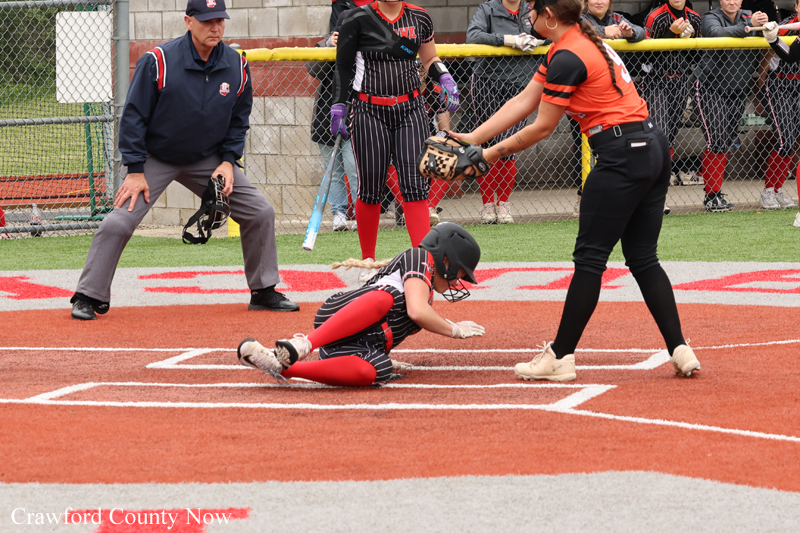 Softball runner slides headfirst into home plate as catcher in black-red gear reaches to tag, with an umpire watching nearby.