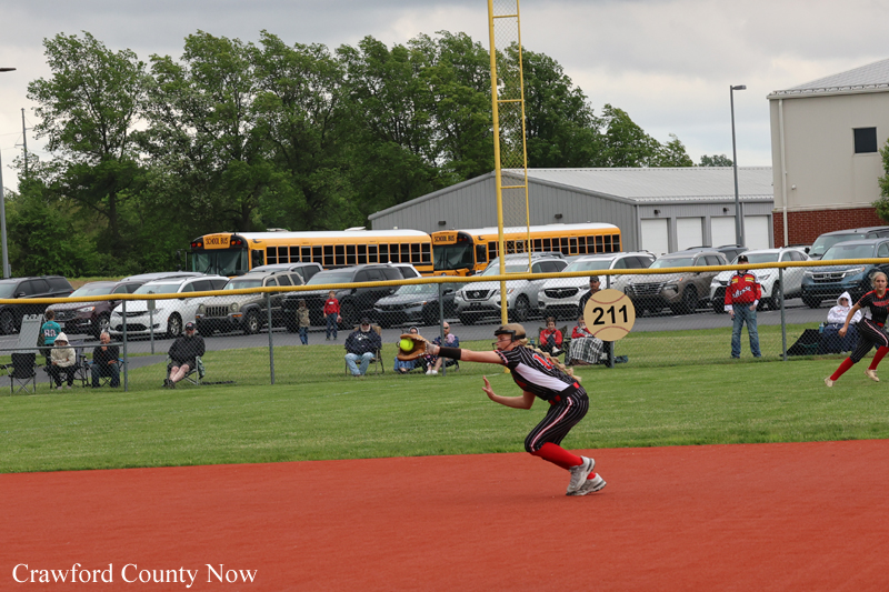 Softball pitcher in a striped uniform throws on an orange dirt infield, with spectators, a chain-link fence, and buses in the background.