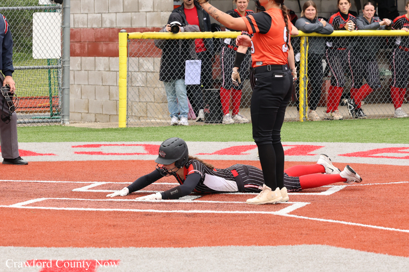 Female softball player in a black uniform slides headfirst into home plate on an orange infield as fans watch from behind the fence.