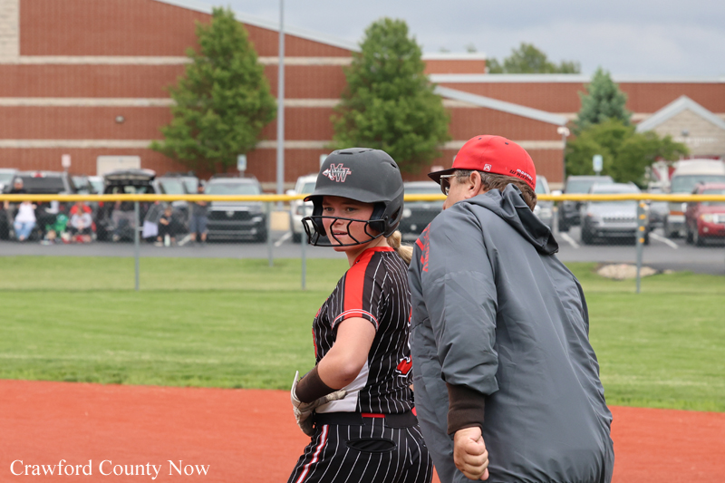Young female softball player in pinstriped uniform with helmet, standing beside a coach on a red infield during practice.