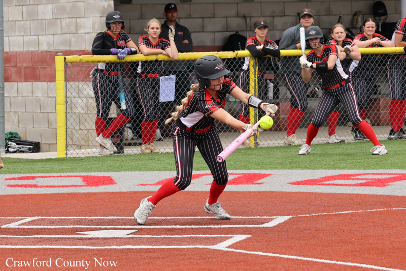 Young softball player in a black-and-red uniform swings at a pitch on a dirt field as teammates watch from the dugout behind a chain-link fence and yellow railing.