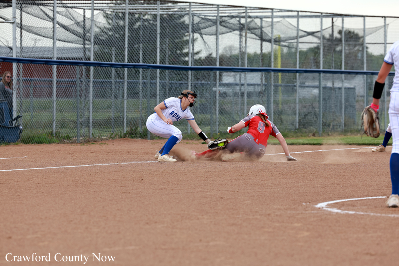 Softball infielder in white tags a sliding baserunner in red as dust sprays near second base. The chain-link fence and field are in the background.