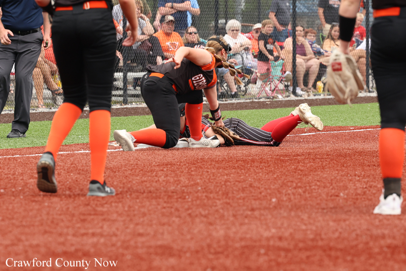 A softball player slides into home plate as a fielder reaches to tag, while teammates and spectators watch from behind the fence.