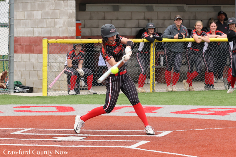 Softball batter in red-and-black uniform swings at a pitched ball on a dirt field, teammates watching from the dugout behind the fence.