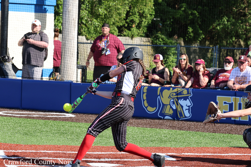 Softball batter in a black pinstripe uniform and red socks swings at a bright green ball, catcher ready nearby, with spectators watching behind the fence.