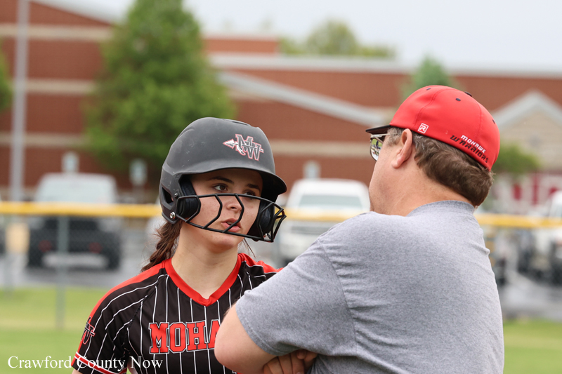 Female softball player in helmet and striped jersey talks with a coach on a softball field.