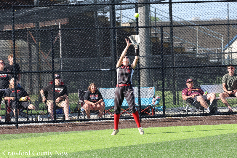 Female softball player in pinstripe uniform catching a high pop fly near the outfield fence, ball in the air above her glove, red socks and beige cleats visible, spectators seated behind the chain-link fence.