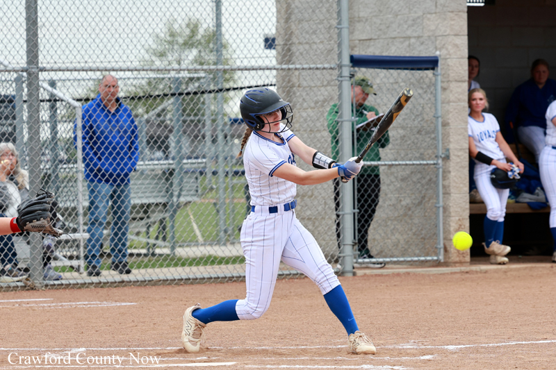 Female softball batter in a white pinstripe uniform swings at a pitch, helmet on, with dugout and fence in background.