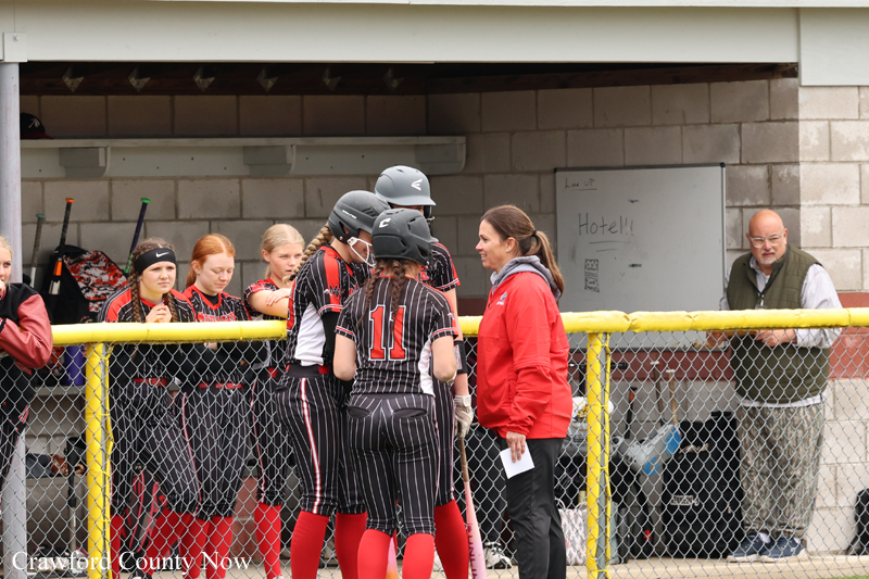 Softball players in black pinstripe uniforms gather in a huddle by a yellow fence while a coach in a red jacket talks to them.