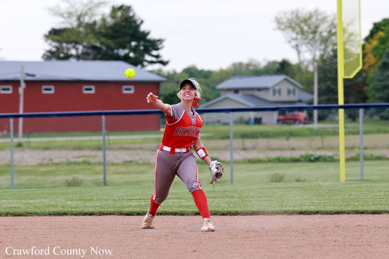 Female softball pitcher in a red uniform throwing a pitch on a dirt infield.