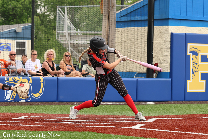 Female softball player in a helmet swings a pink bat during a game on a brown infield, with spectators behind the fence.