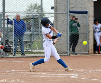 Female softball batter in white-and-blue uniform swings at a pitch during a game, ball visible in flight near her bat, with spectators behind a chain-link fence.