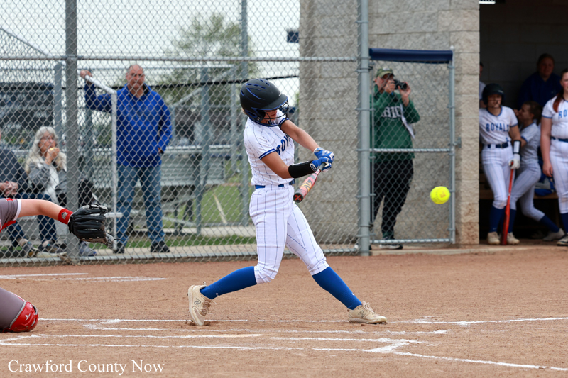 Female softball batter in white-and-blue uniform swings at a pitch during a game, ball visible in flight near her bat, with spectators behind a chain-link fence.